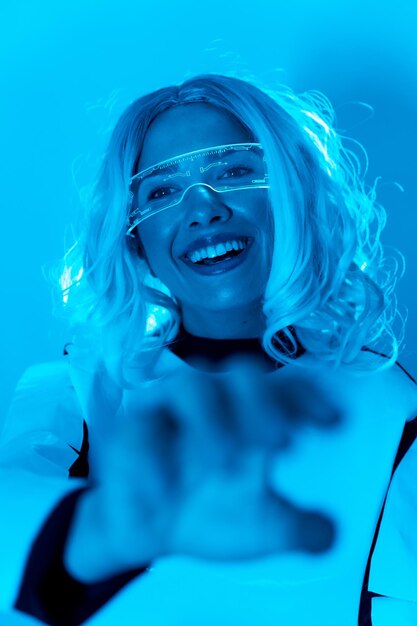 Foto futuristisch concept een jonge vrouw met verlichte virtual reality-bril op een blauwe achtergrond die haar hand reikt