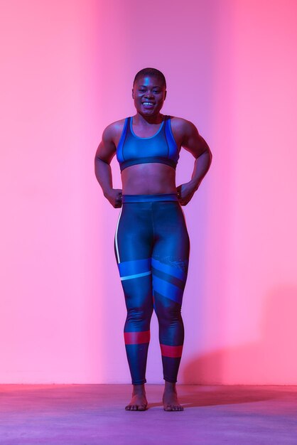 Fysieke activiteit Verticaal portret van de volledige lengte van het lichaam Positieve jonge zwarte vrouw in sporttop BH en legging poseren in de studio Kijken naar camera geïsoleerd op neon roze studio achtergrond Banner