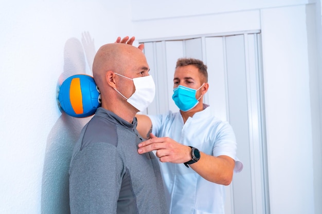 Fysiotherapeut met masker die cliënt helpt om de nek te strekken met een haar op de muur Fysiotherapie met beschermende maatregelen in de Coronavirus pandemie COVID19 Osteopathie sport quiromassage