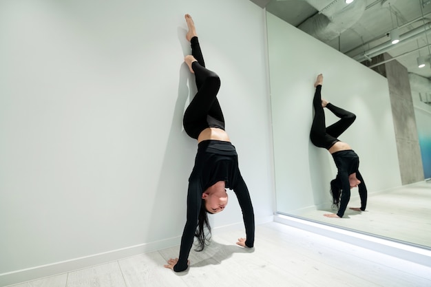 Ga in de buurt van de spiegel staan. Donkerharige actieve vrouw die zich goed voelt terwijl ze een armstand bij de spiegel doet