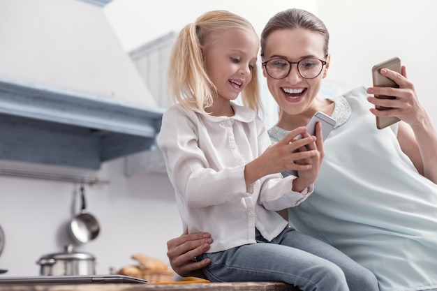 ガジェットの機能。笑いながら画面を見て、電話を持っている女の子に触発された嬉しい陽気な母と娘