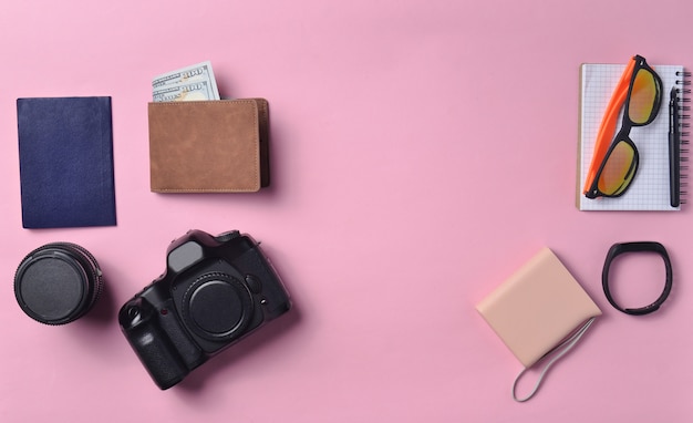 Gadgets and accessories layout on pink pastel background. Photographic equipment, purse with dollars, smart clock, smartphone, notebook, sunglasses, passport, power bank.