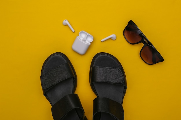 Gadgets and women's accessories on  yellow background. Sandals, smart wireless headphones and sunglasses. Top view. Flat lay