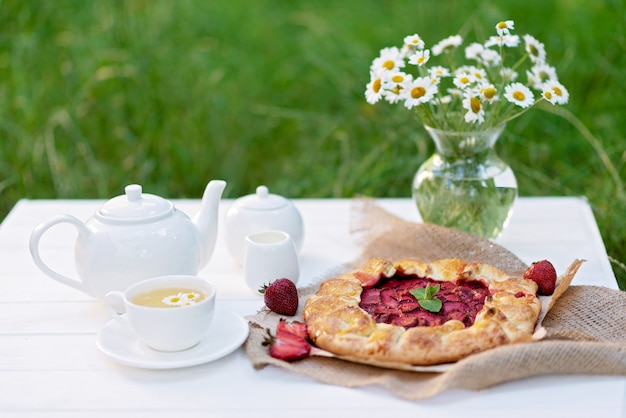 galette aardbeientaart, een kopje kruidenthee, een theepot en een vaas met een boeket madeliefjebloemen.