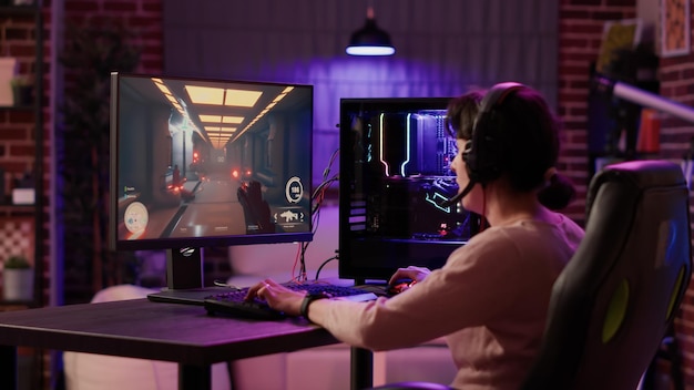 Gamer girl using pc setup playing multiplayer first person shooter talking on headset while explaining gameplay to subscribers. Woman streaming online action game while sitting in gaming chair.