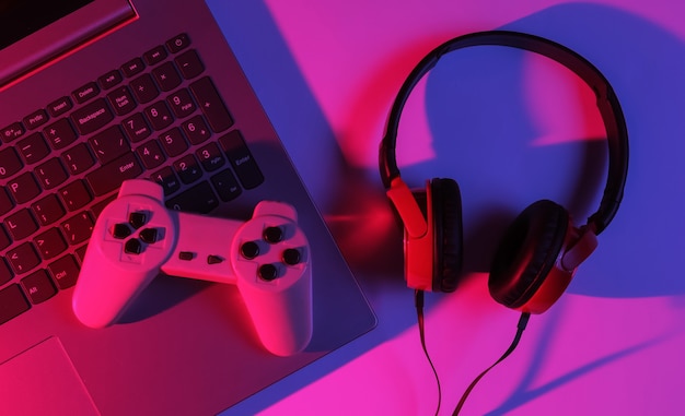 Gaming concept. Gaming equipment. Laptop with gamepad, headphones. Neon red-blue gradient light. Top view