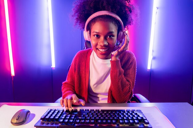 ゲームストリーマーのアフリカの女の子がオンラインで遊んでおり、ネオンカラーの照明室でヘッドフォンを着用してEスポーツの熟練チームと戦っています他のプレイヤーと話し、競合他社に勝つための戦略を計画しています