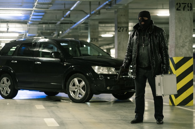 Gangster in zwarte jas en bivakmuts met pistool en koffer terwijl staande op de achtergrond van de auto op de parkeerplaats