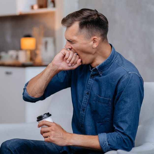 Gapende jonge man met een kopje koffie zittend in een stoel thuisleven