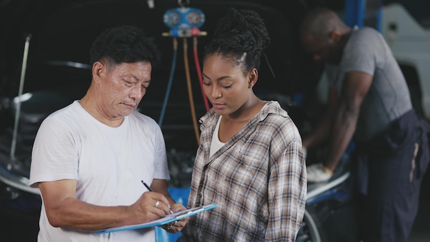 Garage businessman talking explain to car owner about check list service price detail with engine mechanic working in background