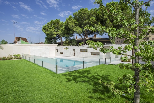 Garden of a detached house with grass and trees and a pool with a glass fence