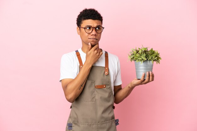 疑いと思考を持っているピンクの背景に分離された植物を保持している庭師アフリカ人