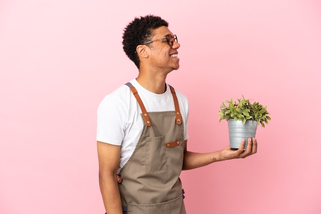 Садовник африканский мужчина держит растение, изолированное на розовом фоне, смеясь в боковом положении