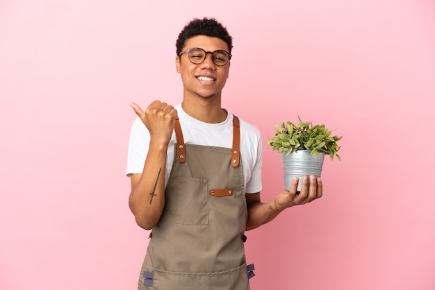 製品を提示する側を指しているピンクの背景に分離された植物を保持している庭師アフリカ人