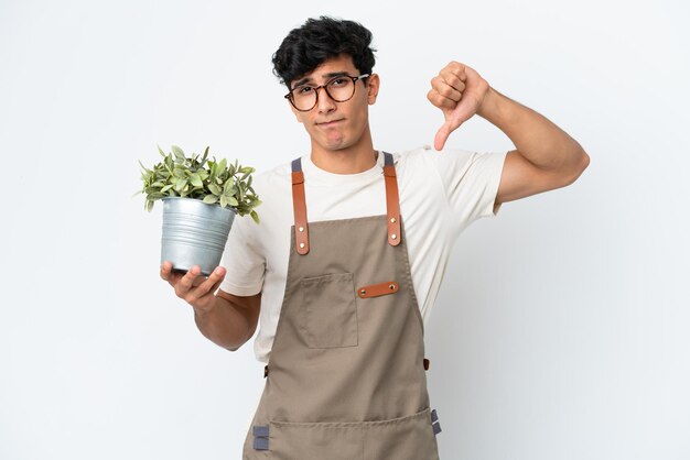 Gardener Argentinian man holding a plant isolated on white background showing thumb down with negative expression