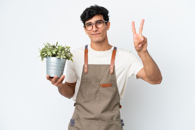 笑顔と勝利のサインを示す白い背景で隔離の植物を保持している庭師アルゼンチン人