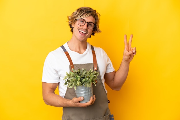 사진 웃 고 승리 기호를 보여주는 노란색 벽에 고립 된 식물을 들고 정원사 금발 남자