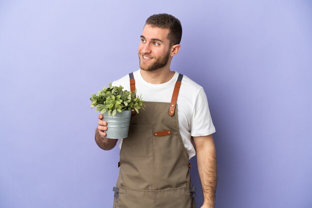 Gardener caucasian man holding a plant isolated on yellow looking to the side and smiling