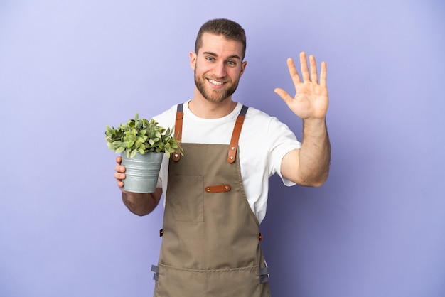 幸せな表情で手で敬礼黄色の壁に分離された植物を保持している庭師の白人男性