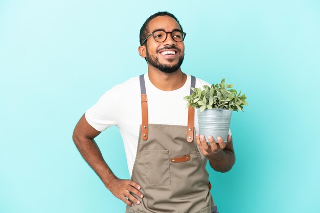 腰に腕と笑顔でポーズをとって青い背景で隔離の植物を保持している庭師ラテン男