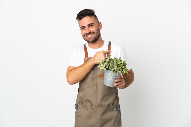 Gardener man holding a plant over isolated white background pointing to the side to present a product