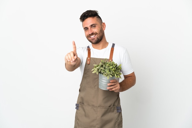 Uomo del giardiniere che tiene una pianta sopra fondo bianco isolato che mostra e che alza un dito