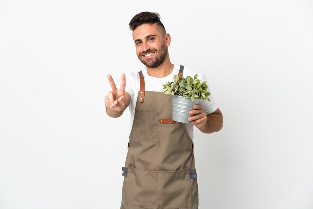 Uomo del giardiniere che tiene una pianta sopra fondo bianco isolato che sorride e che mostra il segno di vittoria