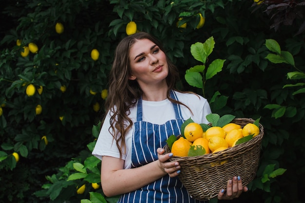 A gardener in a striped blue apron stands in the garden holding a basket of lemons