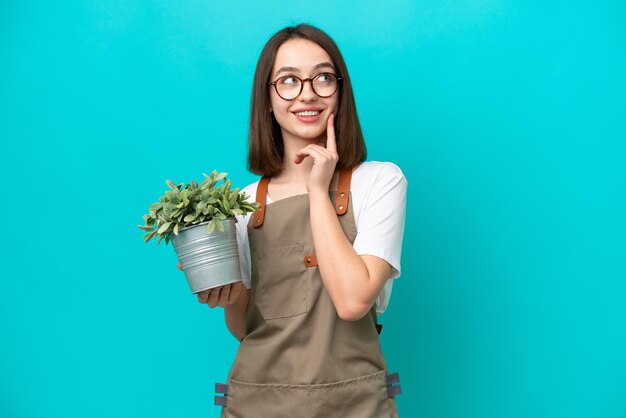 見上げながらアイデアを考えて青い背景で隔離の植物を保持している庭師ウクライナの女性