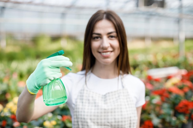 Gardening concept Pretty smiling woman in apron holding sprayer in hand and looking to camera standing in modern greenhouse on the background of plants and flowers Focus on the sprayer