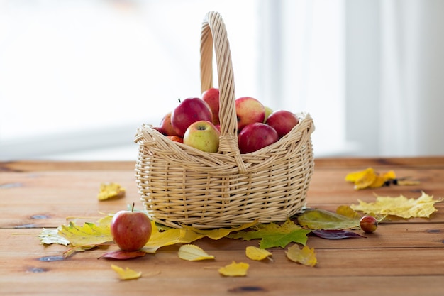 gardening, season, autumn and fruits concept - close up of wicker basket with ripe red apples and leaves on wooden table