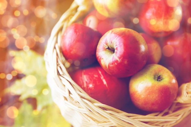 gardening, season, autumn and fruits concept - close up of wicker basket with ripe red apples and leaves on wooden table