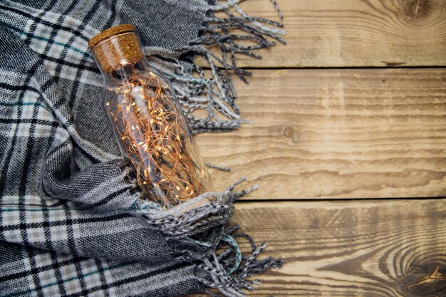 A garland of Christmas lights in a glass bottle.Warm grey scarf or plaid on a wooden background. A postcard with a Christmas holiday mood. Top view with space for text.