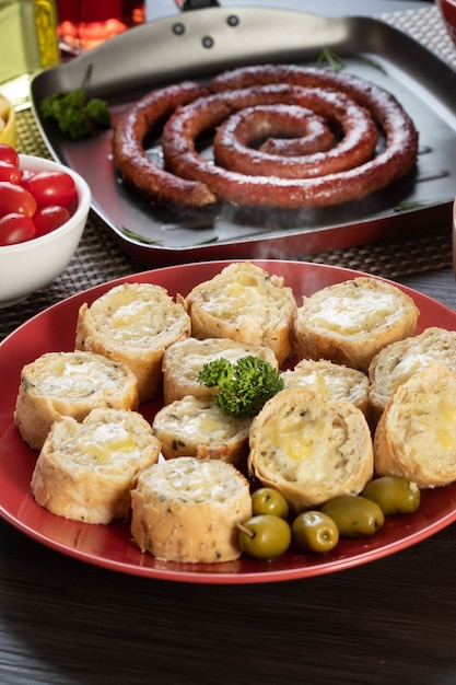사진 소시지, 치즈, 로즈마리, 올리브, 체리 토마토와 함께 바베큐 테이블에 빨간 접시에 마늘 빵.