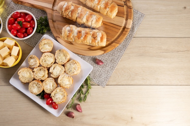 Garlic bread in white square plate on the table with cheese, rosemary, olives and cherry tomatoes. Top view.