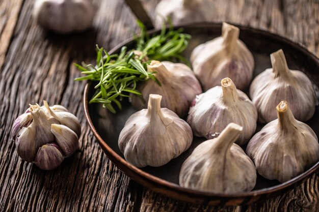 Garlic cloves and bulb in pan with fresh rosemary on old wooden table.