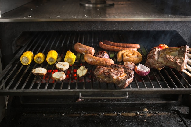 Garlic, onion, corn, tomatoes and meat with sausages on the grill on hot coals with smoke.