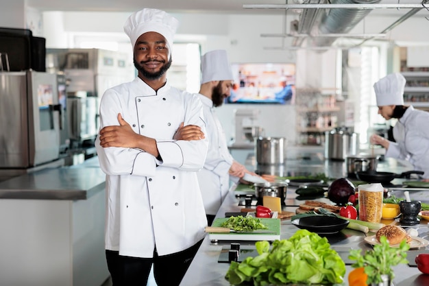 Photo gastronomy expert standing in restaurant professional kitchen with arms crossed while smiling at camera. confident head chef wearing cooking uniform while preparing ingredients for dinner service.