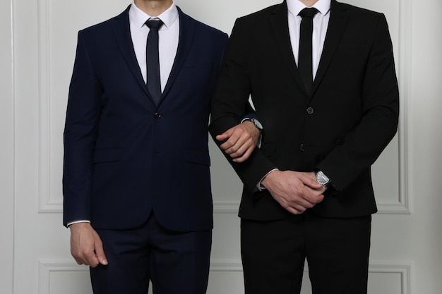 Gay couple in elegant suits standing near white wall closeup