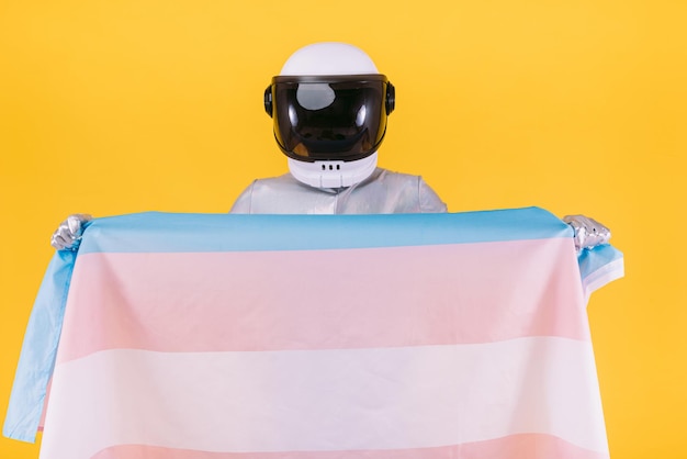 Gay man dressed as an astronaut with a helmet and silver suit holding a flag of the trans community on a yellow background Gay homosexual trans rights and gender pride concept