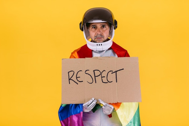 Gay man dressed as an astronaut with a helmet and silver suit with the flag of the lgtbi collective holding a sign that reads 39Respect39 on a yellow background