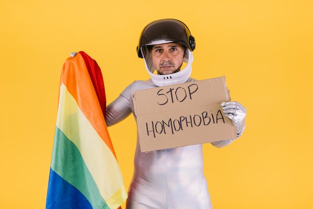 Uomo gay vestito da astronauta con casco e tuta argentata con la bandiera del collettivo lgtbi con in mano un cartello che recita 39stop homophobia39 su sfondo giallo