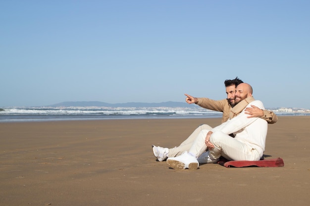 Gay man embracing boyfriend on beach. Middle aged bearded man pointing into distance while talking to lover. Vacations concept