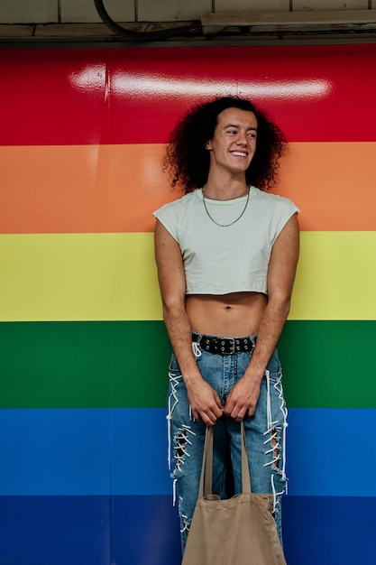 Foto uomo gay con i capelli lunghi e ricci che indossa jeans e un top tagliato con una borsa in mano