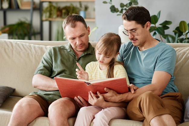 Photo gay parents sitting on sofa and reading a book to their little daughter in the room