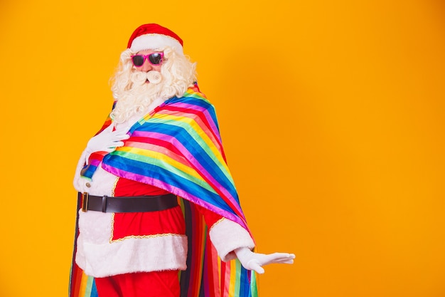 LGBTの旗を持っているゲイのサンタクロース。クリスマスゲイプライドのコンセプト。黄色の背景にゲイサンタクロース。サンタクロースはゲイです