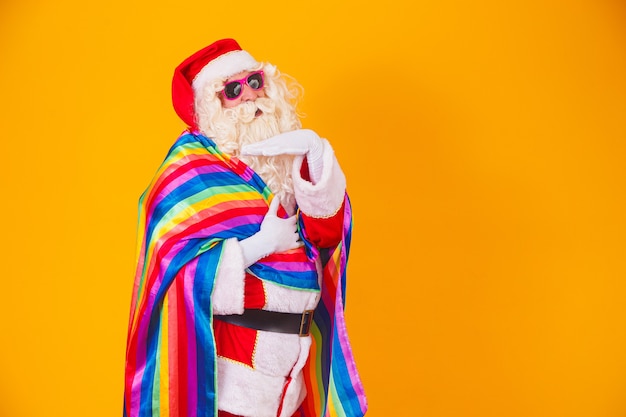 LGBTの旗を持っているゲイのサンタクロース。クリスマスゲイプライドのコンセプト。黄色の背景にゲイサンタクロース。サンタクロースはゲイです