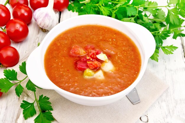 Gazpacho-tomatensoep in een witte kom met groentenlepel op de handdoekknoflook en peterselie op de lichte houten planken als achtergrond