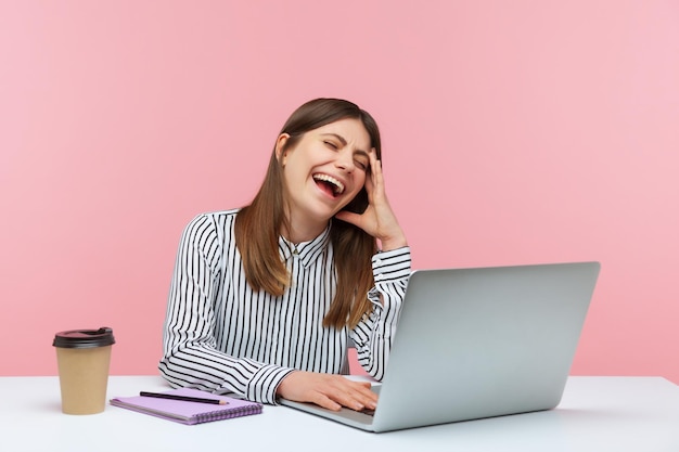 Geamuseerd gelukkige vrouw kantoormedewerker lachen hardop zittend op de werkplek genieten van grappige grap op laptop met pauze thuis kantoor Indoor studio opname geïsoleerd op roze achtergrond