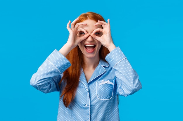 Geamuseerd meisje ziet prachtige promo, wil het proberen. de aantrekkelijke roodharige vrouw in mooie pyjama maakt glazen met vingers, kijkend opgewonden glimlachend en opgewekt, staand in nachtkleding